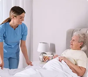 Caregiver tucks in an elderly patient in a bedroom. The patient smiles, next to a nightstand with a lamp.