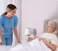 Caregiver tucks in an elderly patient in a bedroom. The patient smiles, next to a nightstand with a lamp.