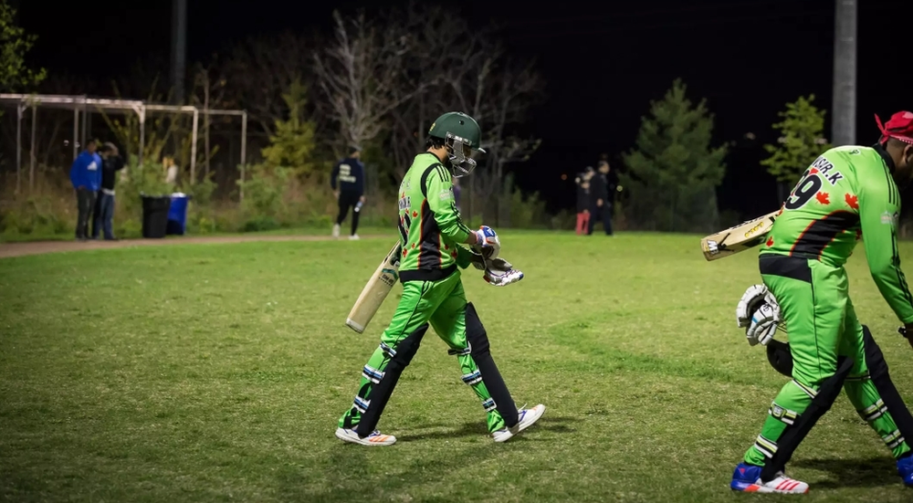 Two cricket players are walking on a field at night.