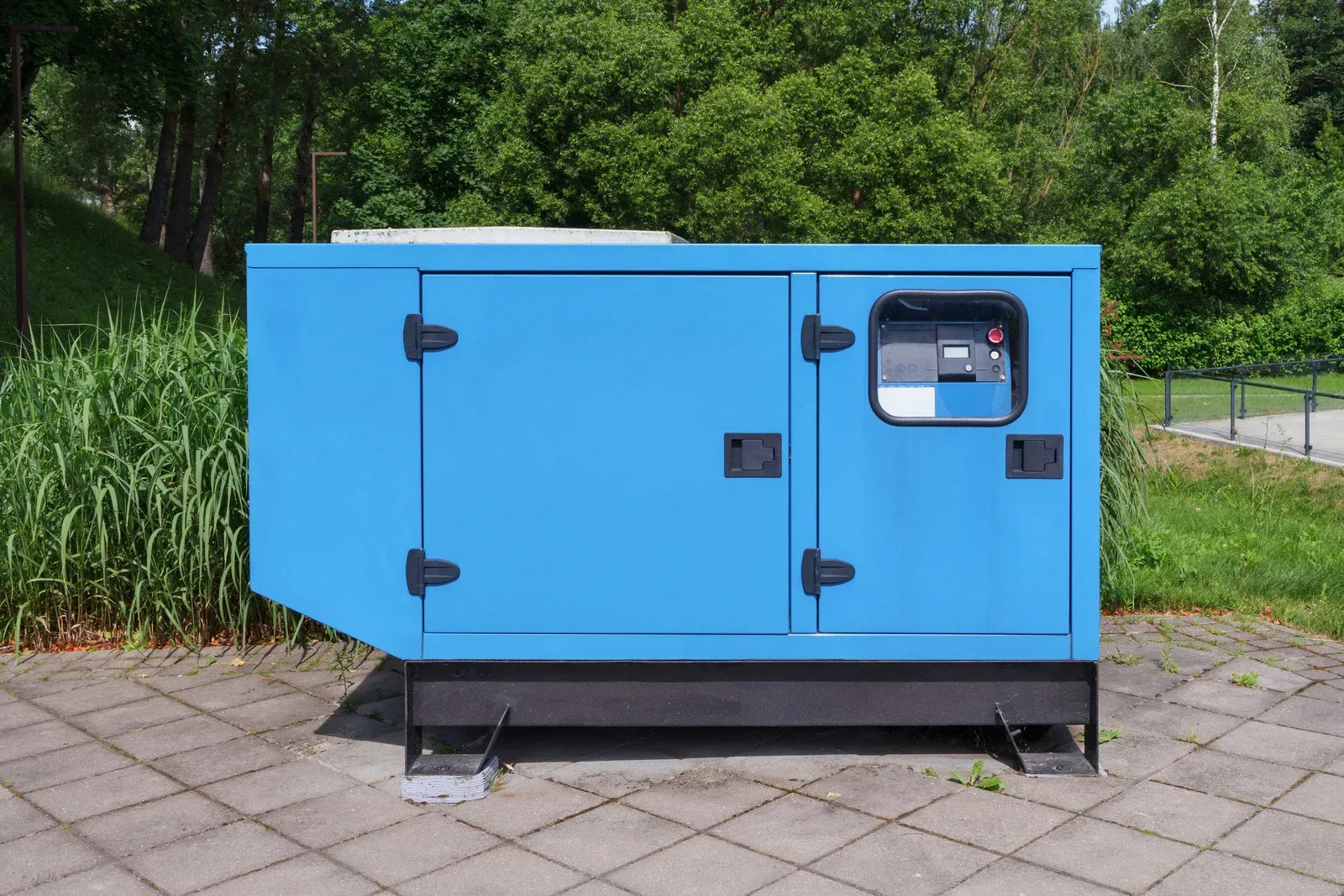 A blue generator is sitting on a sidewalk in a park.
