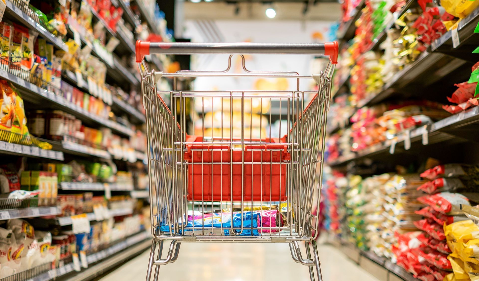 Shopping cart in a grocery store aisle, shelves filled with packaged foods.