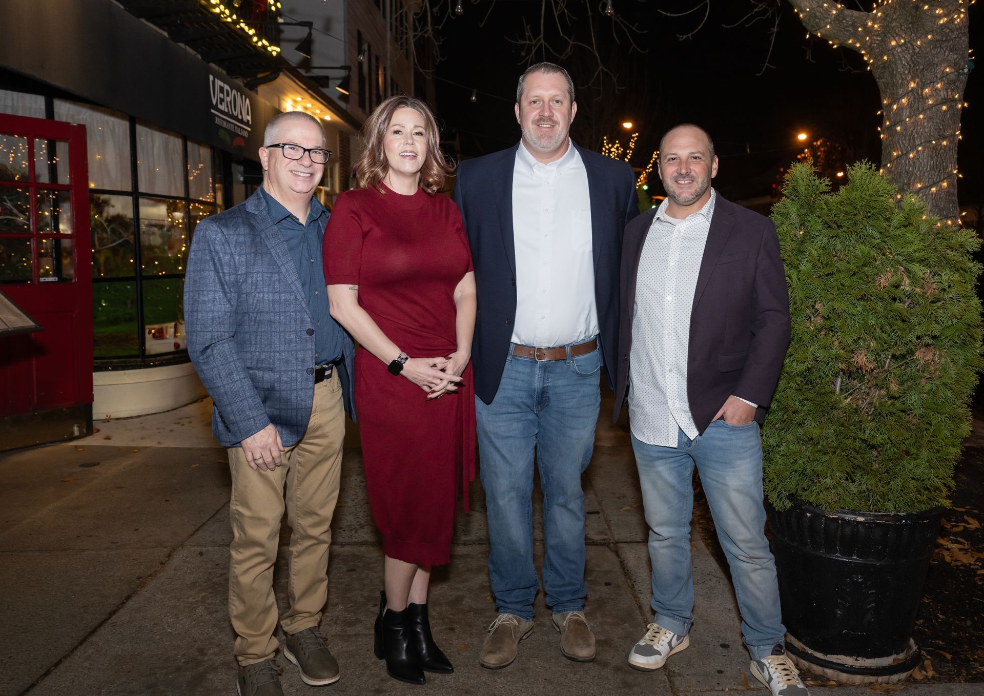 Four people stand outside a restaurant at night. They are smiling, posing for a photo.