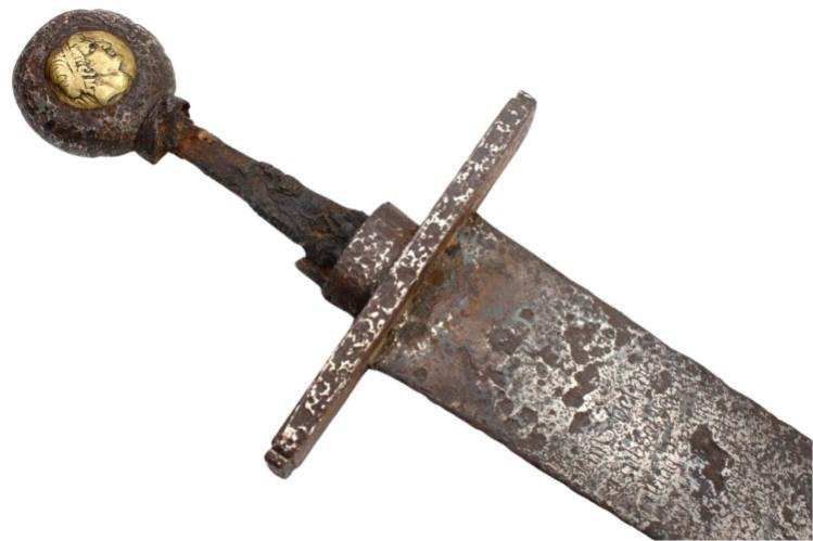 Close-up of an old, rusted sword. The hilt has a circular pommel with gold details.