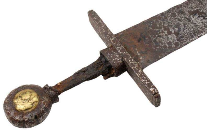 Close-up of a rusted sword hilt and blade, featuring a disc-shaped pommel with a gold inlay.