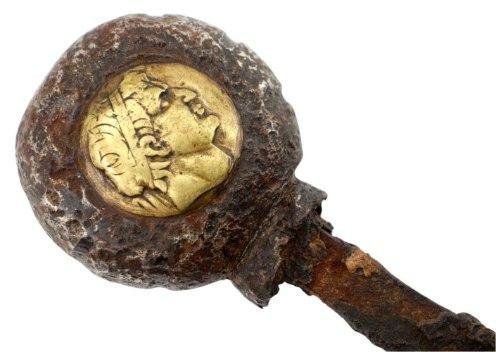 Ancient gold coin with a portrait of a man inside a rusty, rounded frame, attached to a corroded handle.