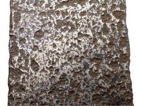 Close-up of a roughly textured, metallic surface, showing a dark brown pitted pattern against a silvery background.