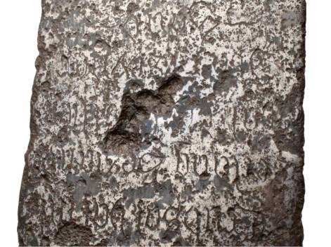 Weathered stone tablet with engraved text, possibly an old tombstone. The surface is gray with a large hole and worn lettering.