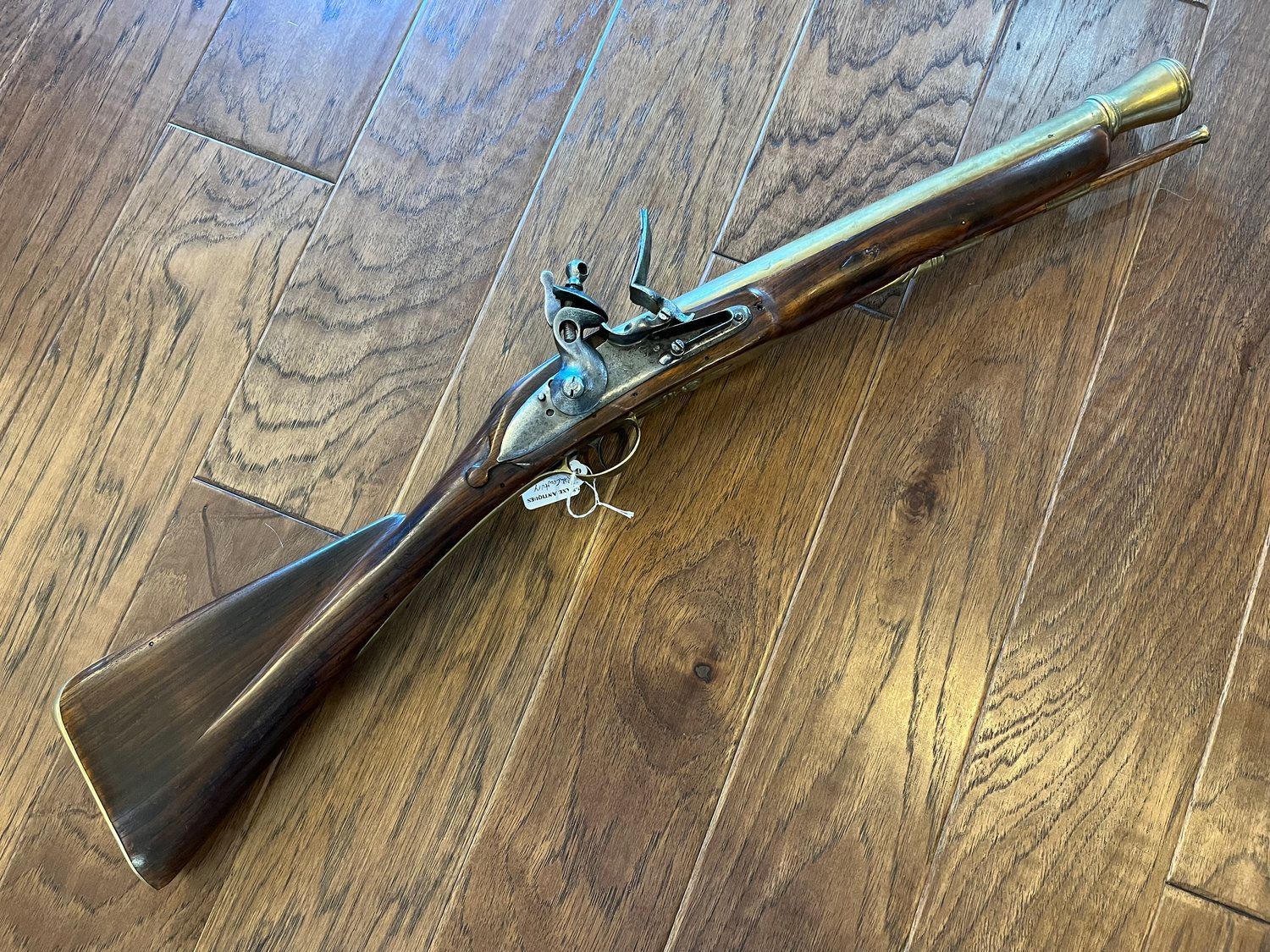 A long, antique flintlock rifle with a wooden stock and brass accents rests on a hardwood floor.