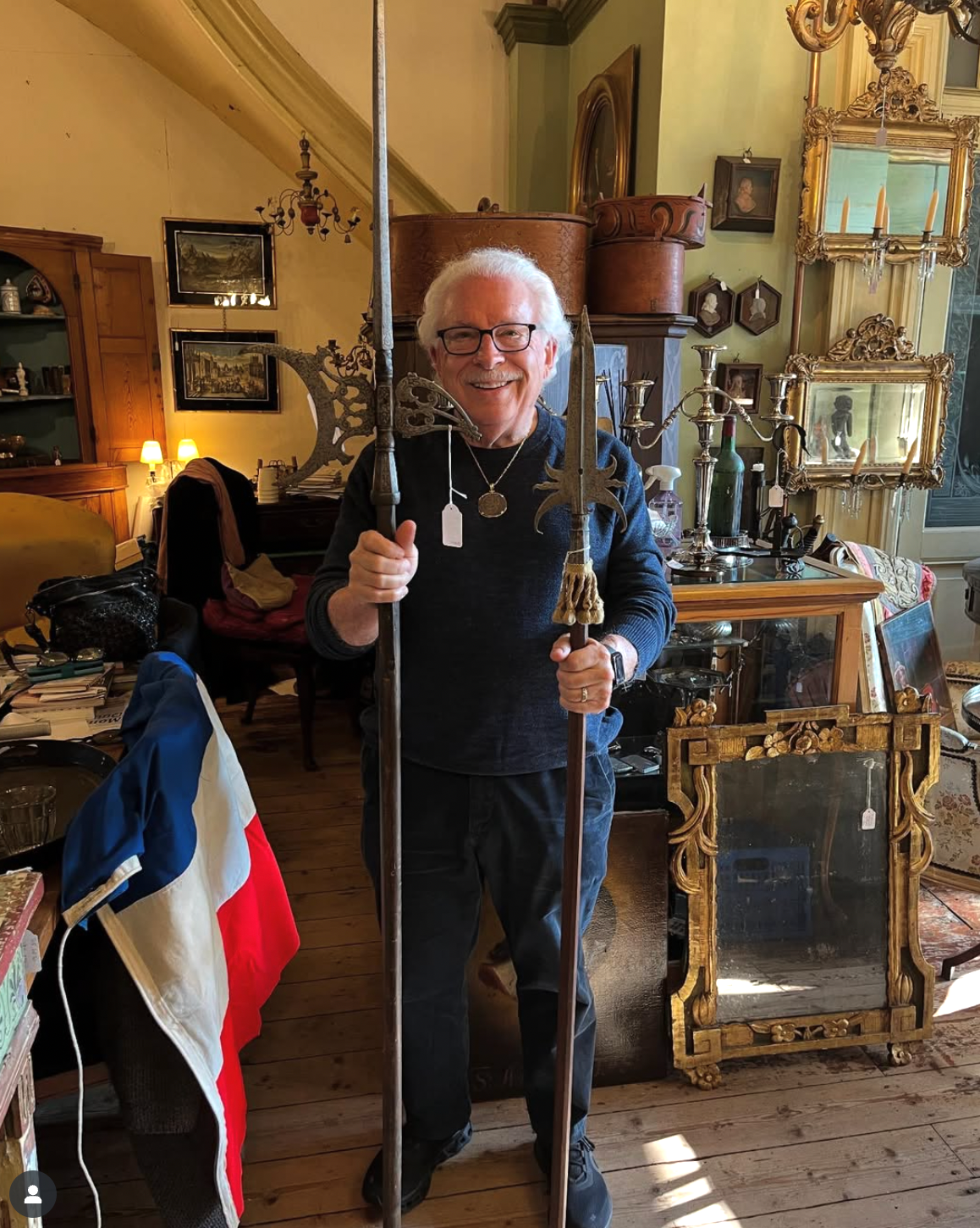 Man holding two spears, smiling in an antique shop with mirrors, furniture, and a French flag.