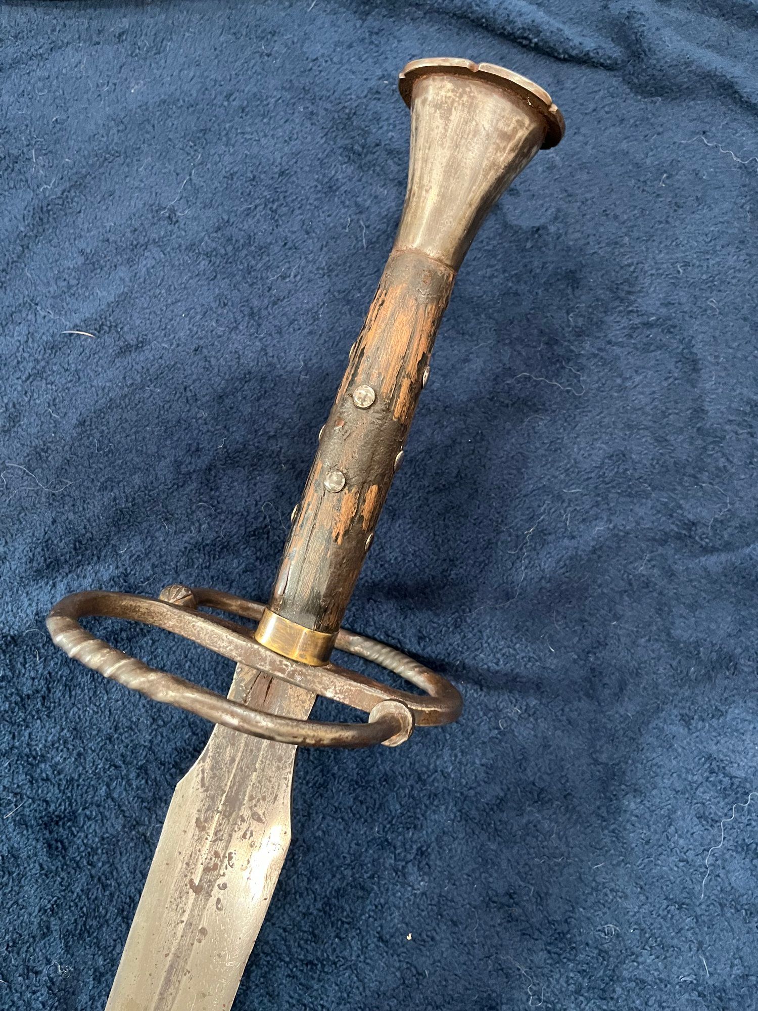 Close-up of an antique sword with a worn wooden handle, circular guard, and faded, corroded blade, resting on a textured blue surface.