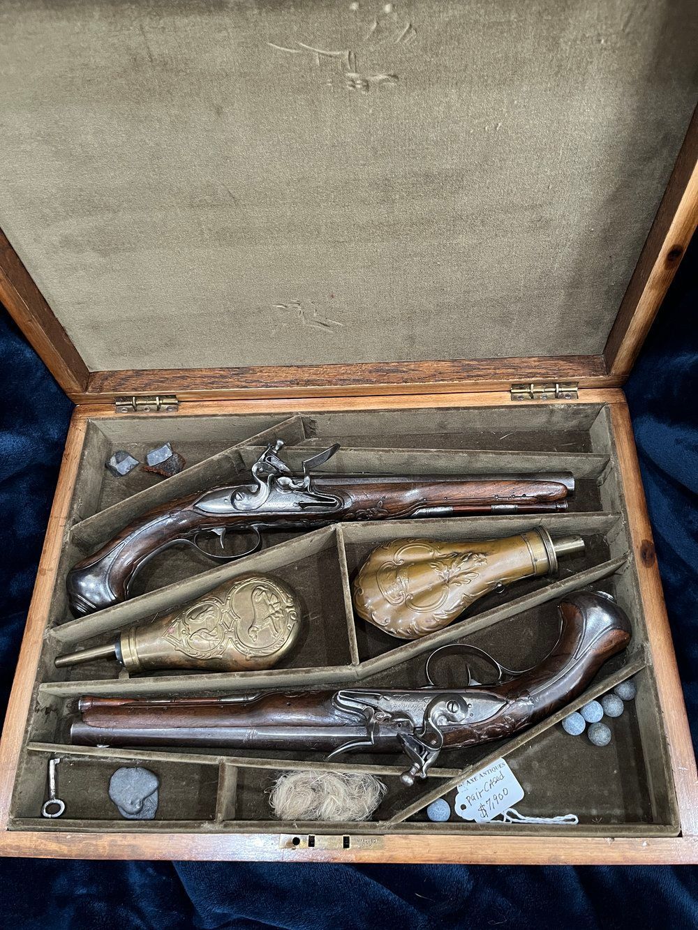Open wooden case holding two antique pistols, a powder flask, a shot bag, and accessories.