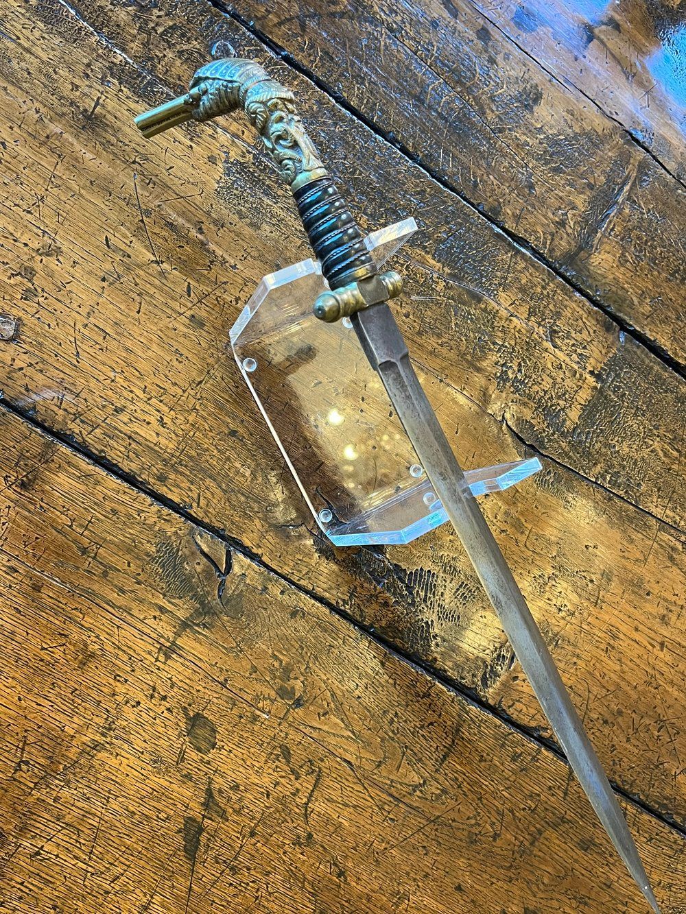 Dagger displayed on a clear stand, set against a wood grain surface. Features ornate hilt and straight blade.