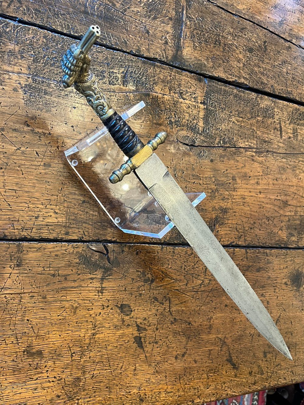 Dagger displayed on a clear stand, set against a wood grain surface. Features ornate hilt and straight blade.