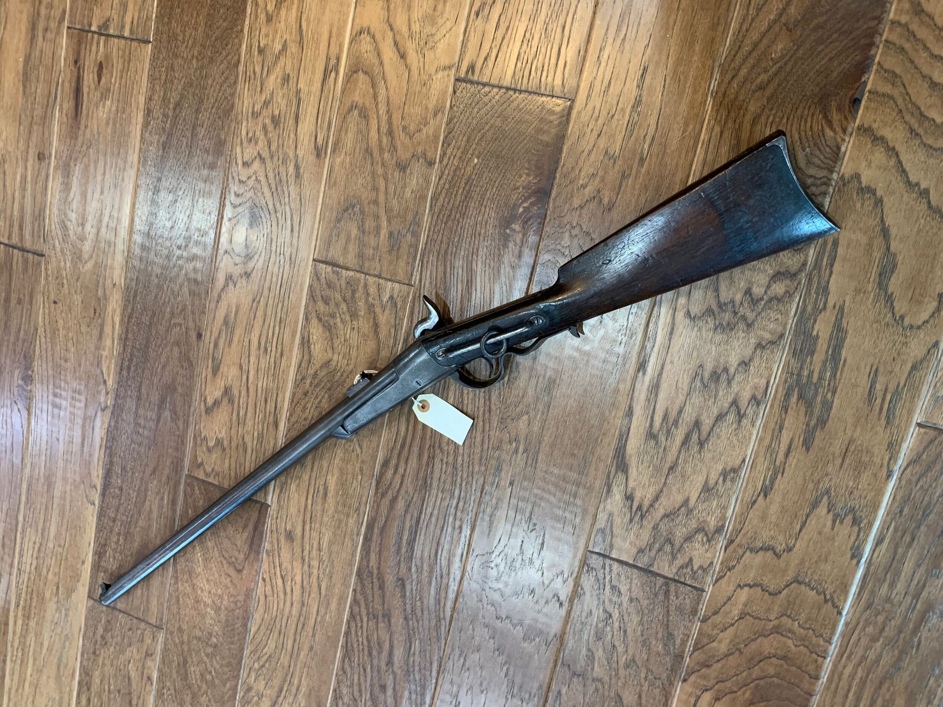 Antique rifle laying on a wooden floor, featuring dark metal and wood. A tag is attached near the trigger.