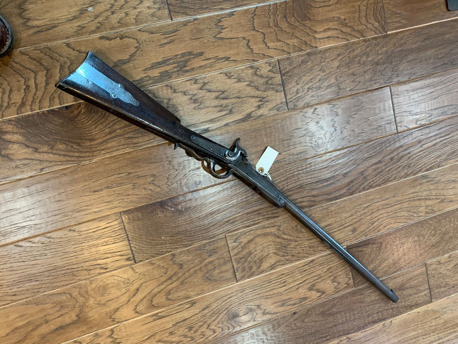 Antique rifle laying on a wooden floor, featuring dark metal and wood. A tag is attached near the trigger.