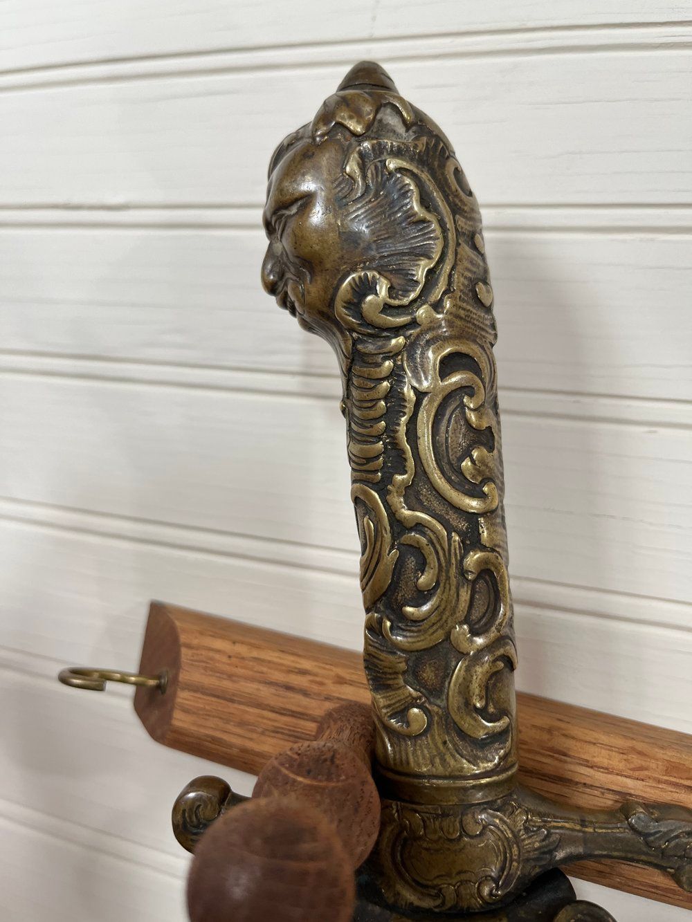 Ornate bronze sword hilt with a face on top and scrollwork details, mounted on a wooden rack.