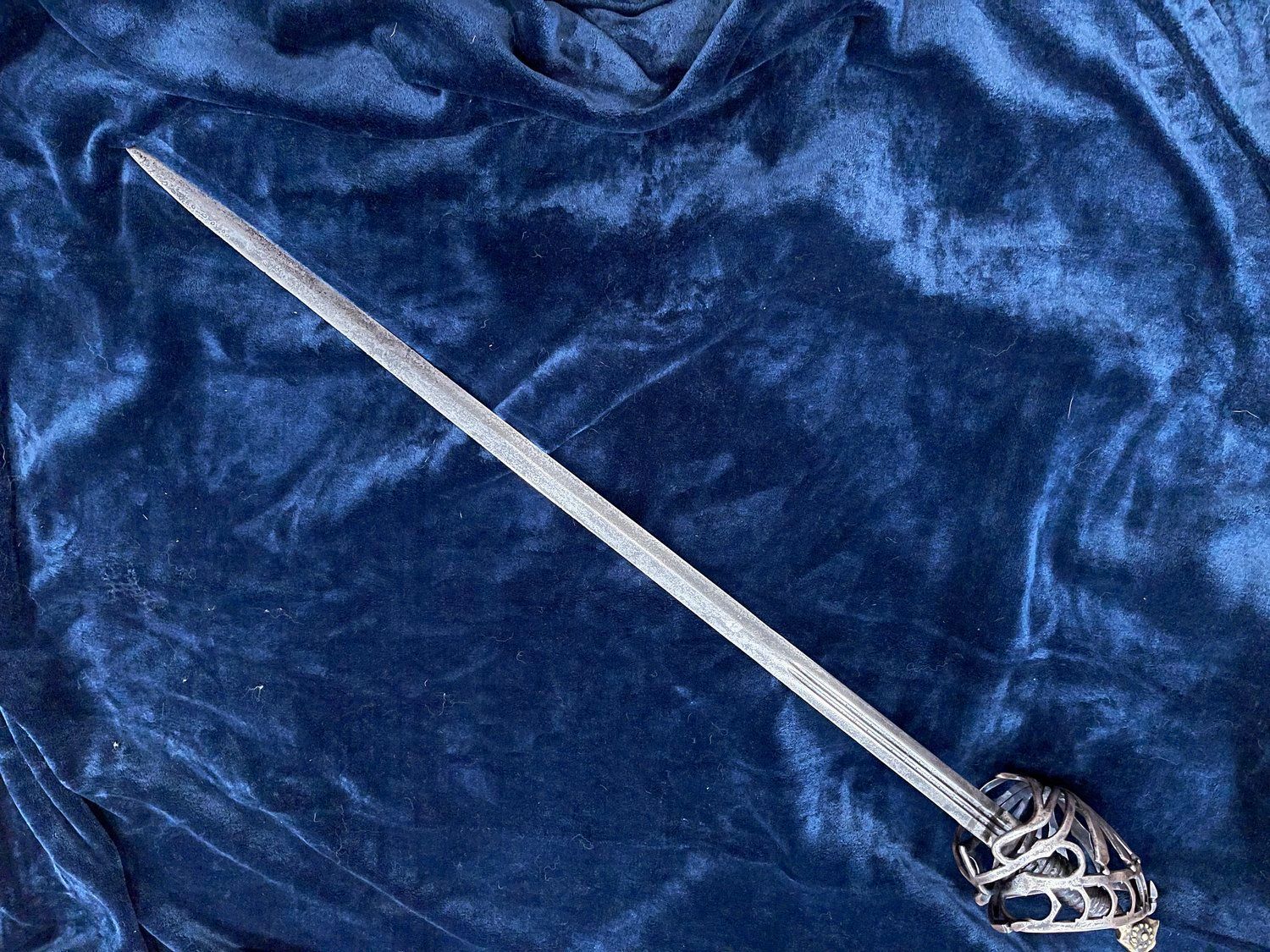 A silver saber with an ornate hilt on a wrinkled blue velvet surface.
