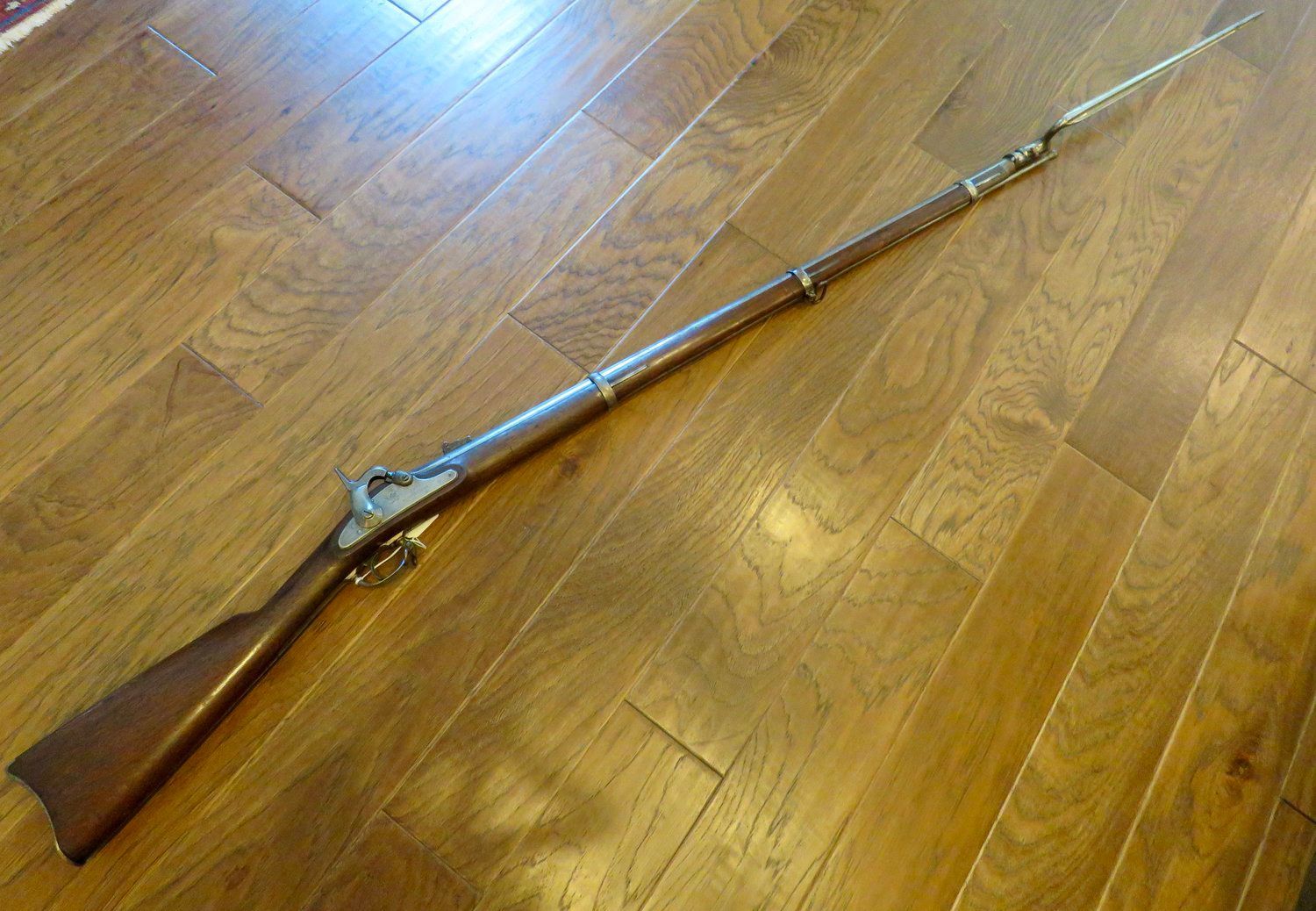 Antique rifle with a bayonet attached, resting on a wooden floor. The rifle is brown and silver, with some visible wear.