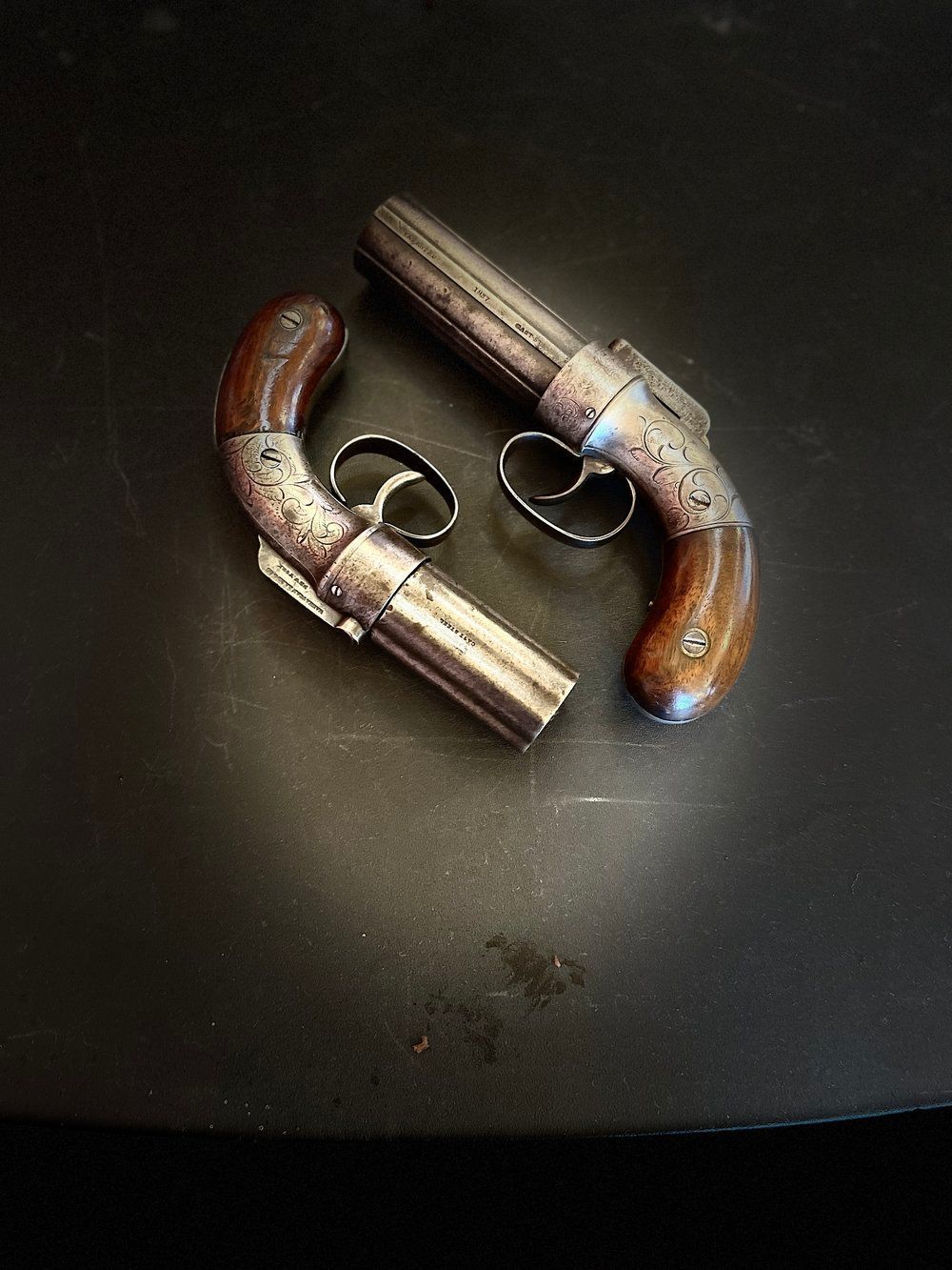 Two antique pocket pistols with wood handles rest on a dark, reflective surface. The guns are silver and show signs of wear.