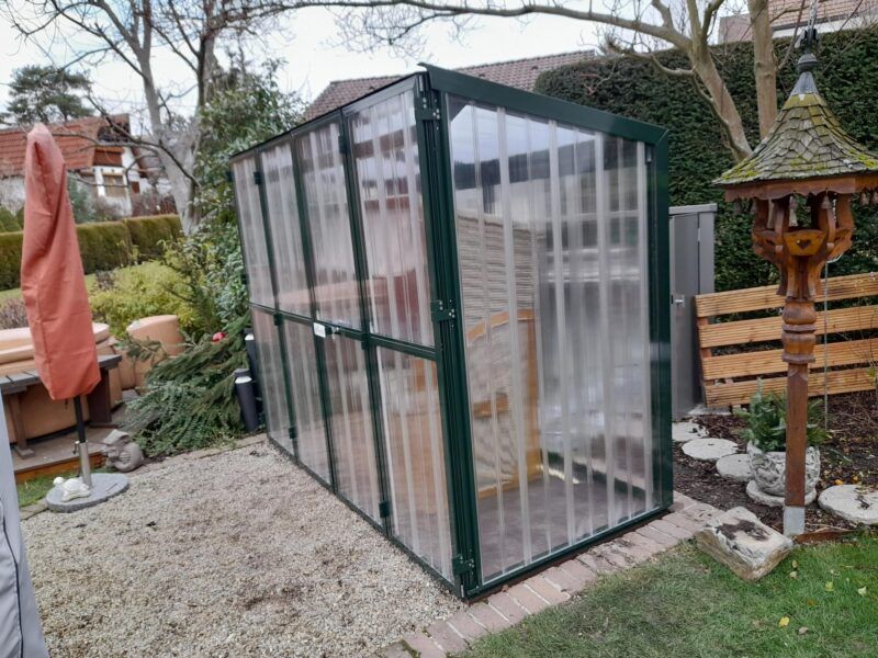 Gewächshaus in Gartenlage, konstruiert aus Wellkunststoff und grünem Metallrahmen.