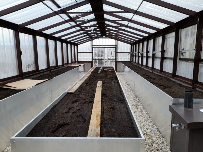 Innenansicht eines Gewächshauses mit erhöhten Metallbeeten, die mit dunkler Erde gefüllt sind, und einem lichtdurchlässigen Dach.