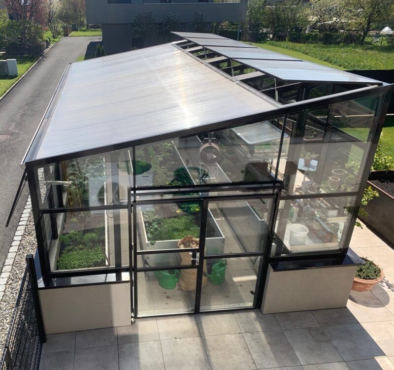 Gewächshaus mit schwarzem Rahmen, Glaswänden und schrägem Dach, in dem sich Pflanzen und Hochbeete befinden.