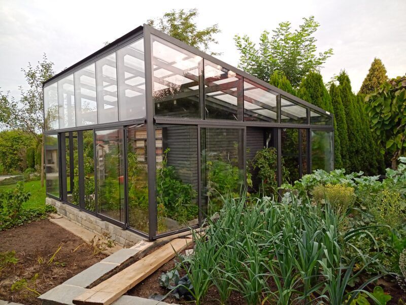 Gewächshaus mit Glaswänden und -dach, umgeben von einem Garten mit verschiedenen Pflanzen.