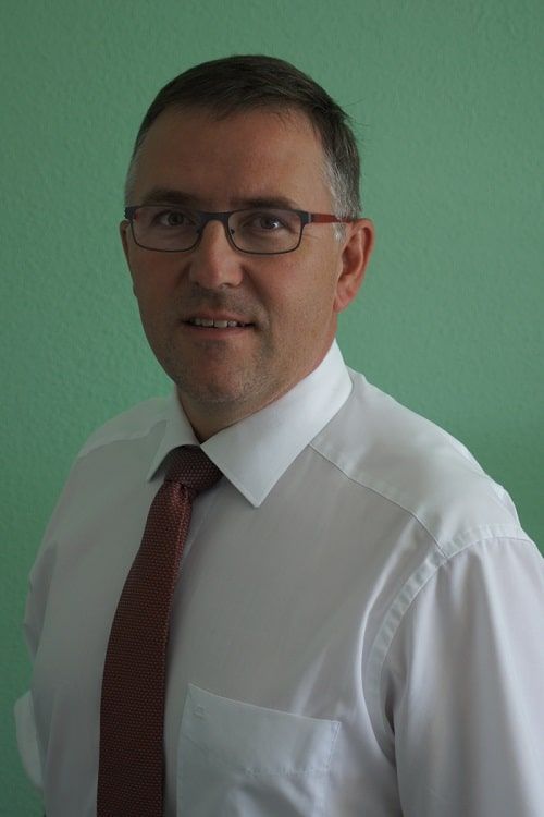 Mann mit Brille, weißem Hemd und Krawatte vor einer grünen Wand.