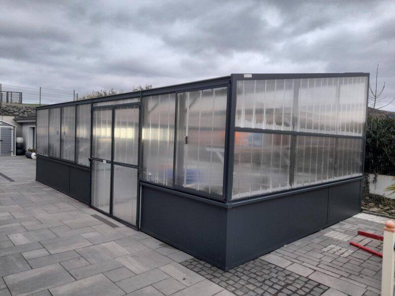 Ein rechteckiges, dunkelgraues Gewächshaus mit durchsichtigen, gewellten Kunststoffplatten und einem bewölkten Himmel im Hintergrund.