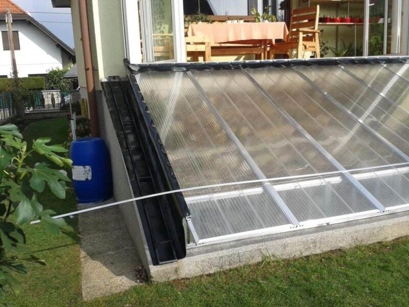 Ein an ein Gebäude angebautes Gewächshaus mit lichtdurchlässigen Dachplatten, einer Dachrinne und einem blauen Wasserfass in der Nähe.