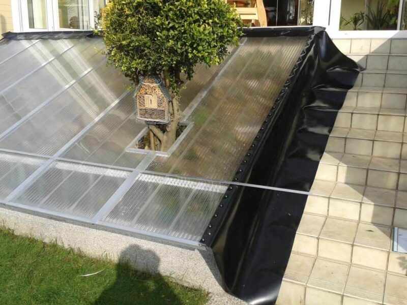 Ein Wintergarten mit transparentem Dach und einem darin wachsenden Baum; schwarze Einfassung an den Stufen.