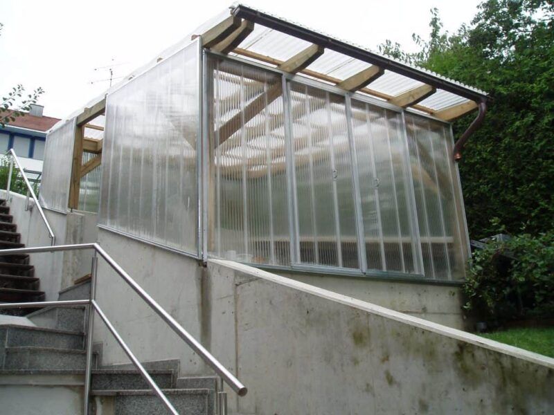Gewächshaus mit transparenten Wänden und Dach neben einer Außentreppe mit Metallgeländer.