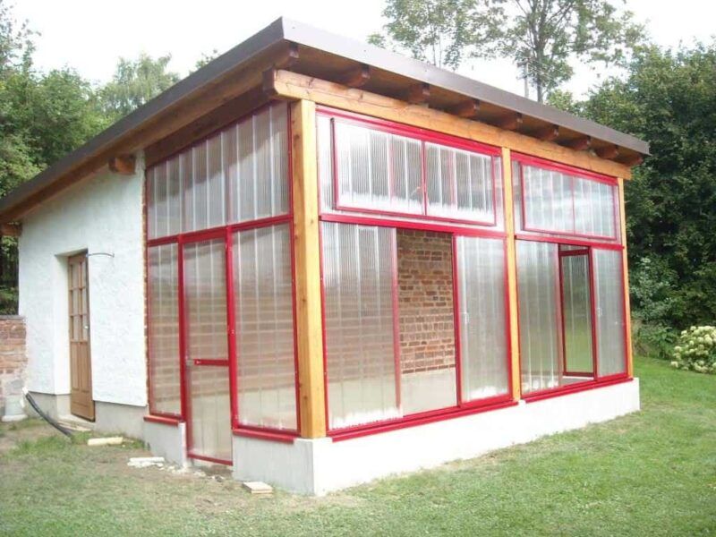 Wintergarten mit rot umrandeten Fenstern aus Wellkunststoff und Holzdach, an ein weißes Gebäude angebaut.