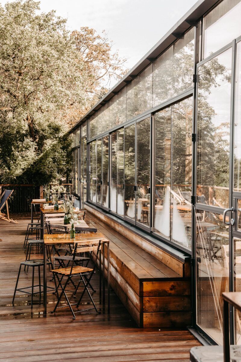 Essbereich im Freien mit verglastem Gebäude, Holzterrasse, Tischen und Bäumen im Hintergrund.