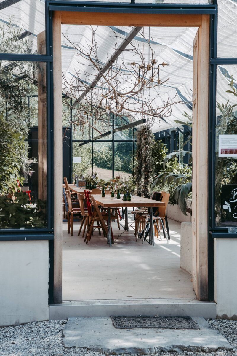 Blick auf einen sonnendurchfluteten Gewächshaus-Essbereich mit Holztisch und Stühlen, Pflanzen und Bäume sind zu sehen.