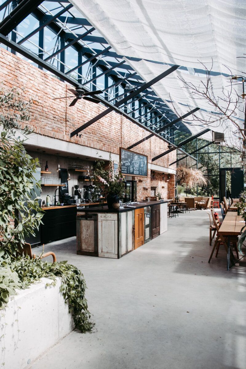 Café mit Glasdach, Backsteinwänden, Bar und Tischen. Pflanzen und natürliches Licht schaffen einen hellen, luftigen Raum.