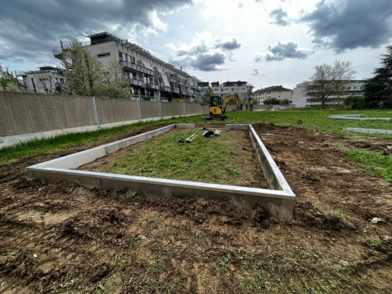 Ein rechteckiges Betonfundament auf einem unbefestigten Feld, im Hintergrund ein kleiner Bagger und Gebäude.