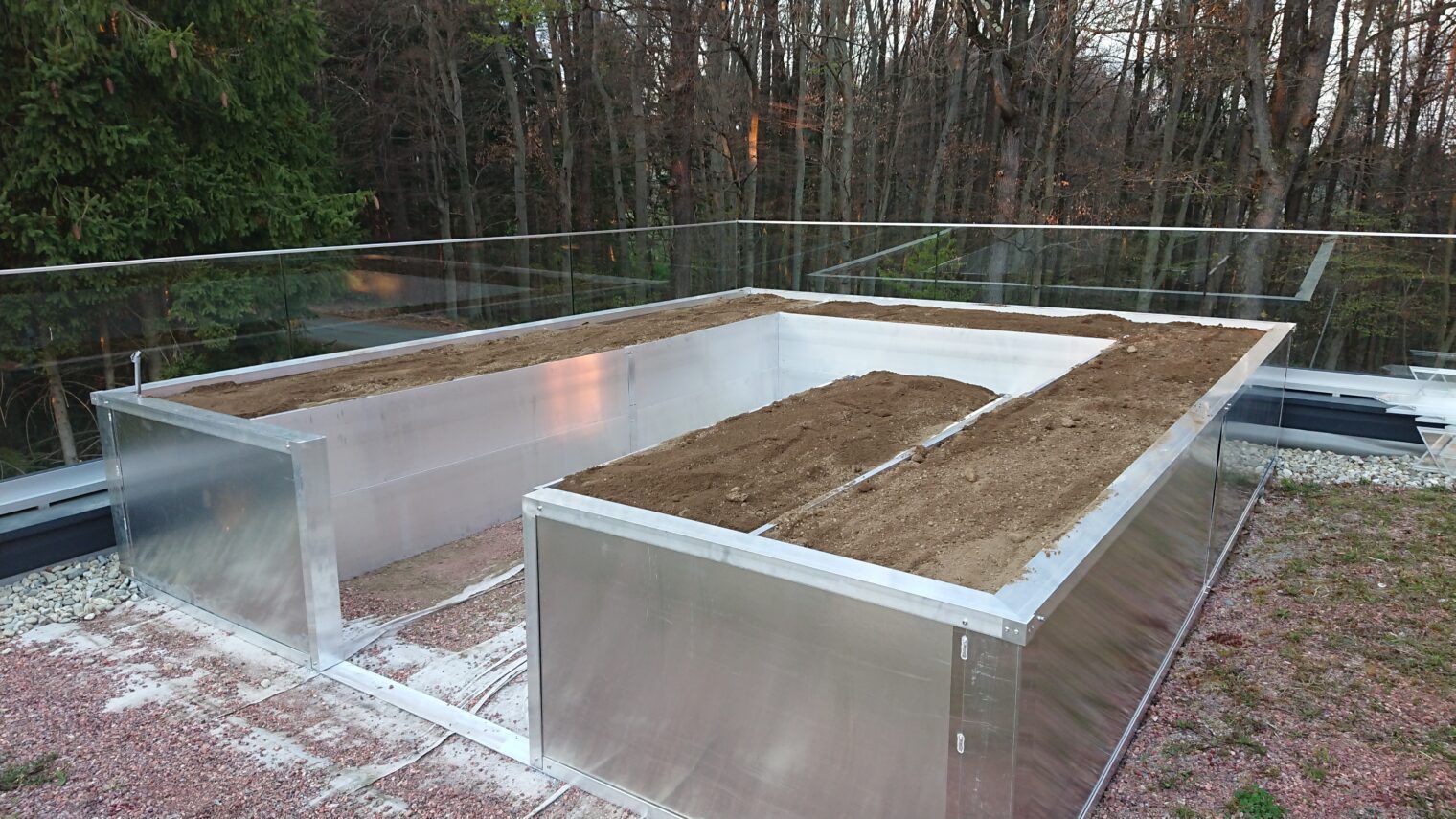Rechteckige Pflanzkästen aus Metall auf einem Dach, gefüllt mit Erde und Pflanzen, umgeben von einem Glasgeländer, im Hintergrund Bäume.