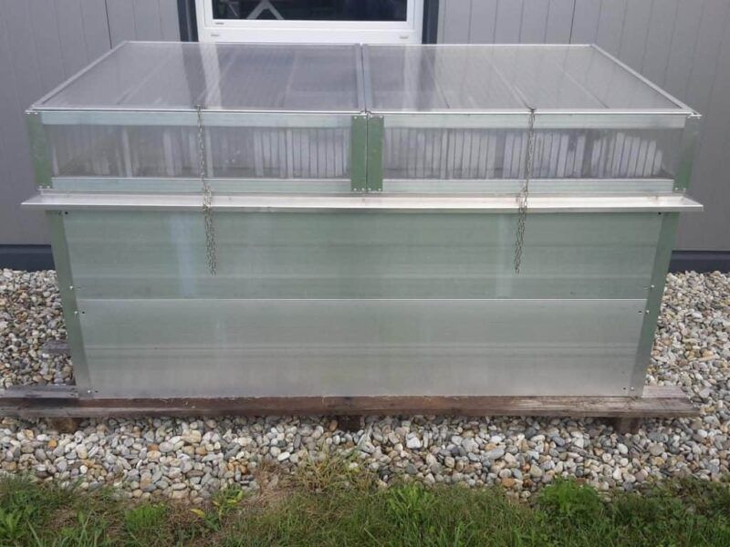 Ein durchsichtiges Kunststoff-Frühbeet auf einem Kiessockel vor einem grauen Gebäude.