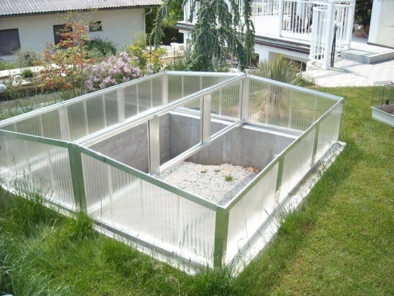 In einem grasbewachsenen Hof steht ein kleines, durchsichtiges Gewächshaus auf einem Betonsockel.
