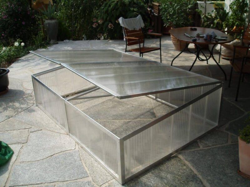 Auf einer Steinterrasse steht ein rechteckiges Frühbeet mit durchsichtigem, klappbarem Deckel.