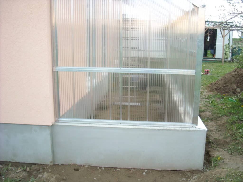 Gewächshaus mit durchsichtigen Kunststoffplatten, die an einem Betonfundament neben einer pfirsichfarbenen Wand befestigt sind.