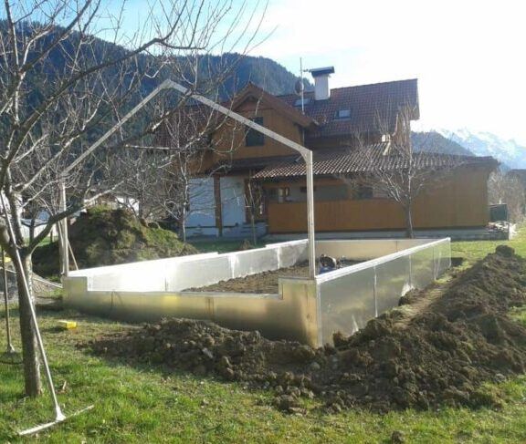 Gewächshausgerüst im Bau in einem Garten mit einem Haus und Bergen im Hintergrund.