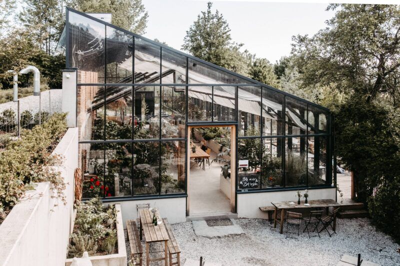 Gewächshaus mit Glaswänden und schrägem Dach, offener Tür, Steinterrasse, Sitzgelegenheiten im Freien.