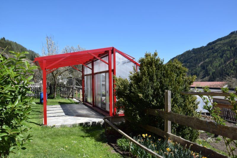 Gewächshaus mit rotem Rahmen und Vordach, eingebettet in einen sonnigen Garten mit Bergen im Hintergrund.