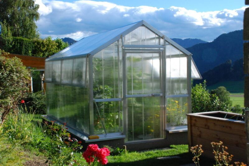 Gewächshaus mit transparenten Wänden und Dach, eingebettet in einen Garten mit Blumen und Bergen im Hintergrund.