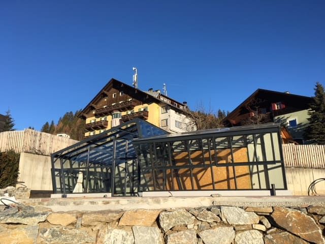 Verglaste Konstruktion mit schwarzen Rahmen vor zwei Holzgebäuden unter blauem Himmel.
