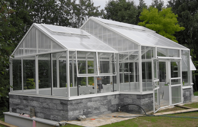 Ein weißgerahmtes Gewächshaus mit transparenten Paneelen steht auf einem Steinfundament und verfügt über eine Tür und mehrere Fenster.
