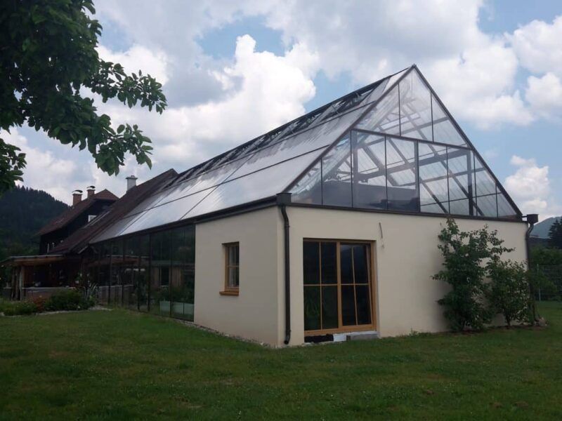 Langer Gewächshausanbau mit Glasdach an einem weißen Gebäude, das auf einem grünen Rasen steht.