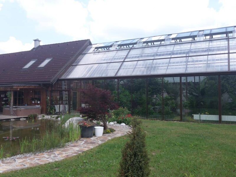 Ein Haus mit einem großen, angebauten Glasgewächshaus, einem Teich und einem grünen Garten.